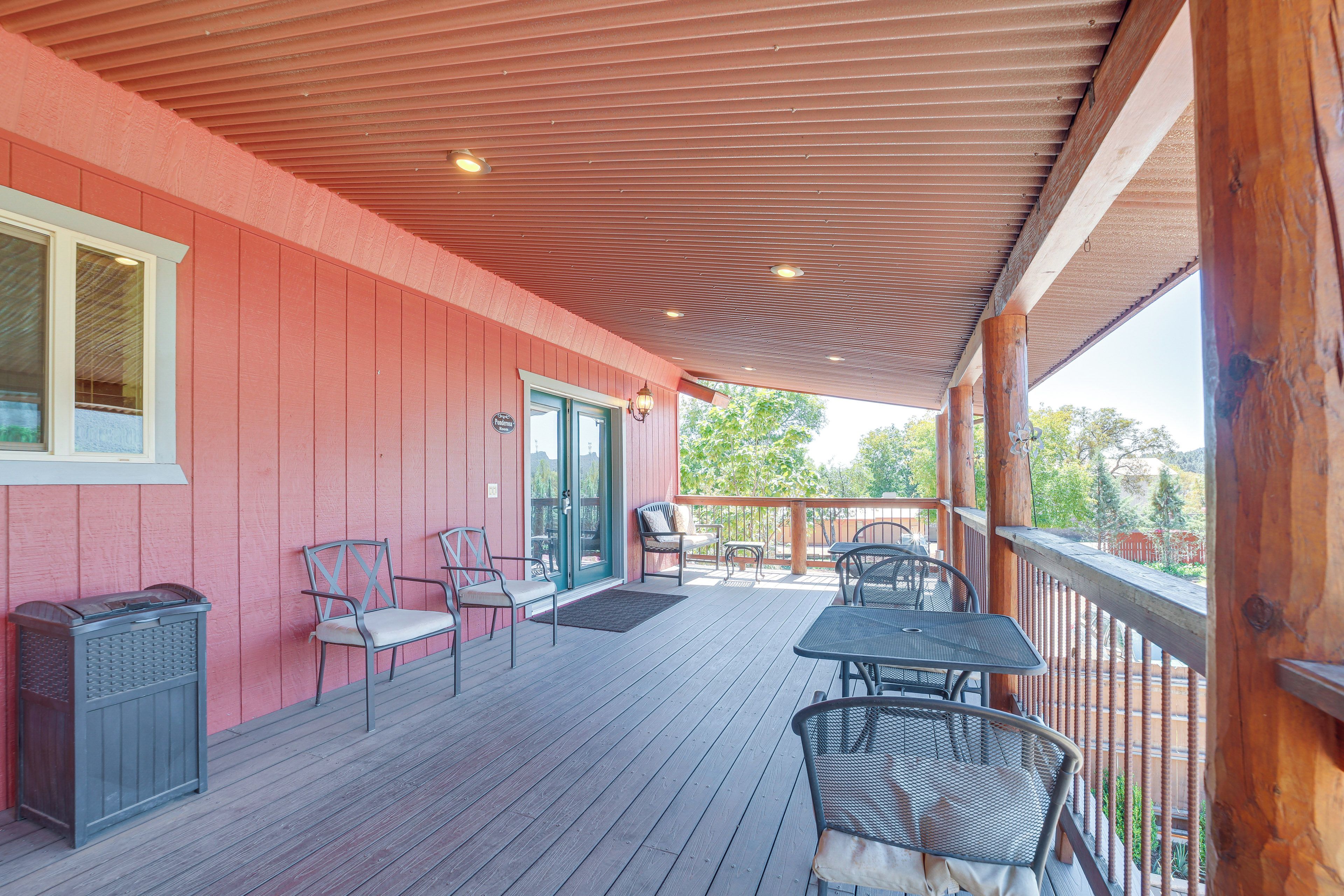A covered patio with wooden flooring, metal chairs, a table, and a trash can.