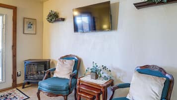 Una sala de estar con dos sillones tapizados en azul, una mesita de madera con un florero, un televisor de pantalla plana montado en la pared y un antiguo estufa de leña.