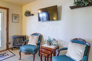 Una sala de estar con dos sillones tapizados en azul, una mesita de madera con un florero, un televisor de pantalla plana montado en la pared y un antiguo estufa de leña.