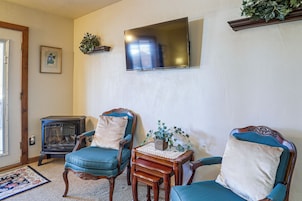A living room with two blue upholstered chairs, a small wooden table with a vase, a wall-mounted flat-screen TV, and a vintage wood-burning stove.