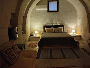 Quarto aconchegante e com iluminação suave, com cama, sofá e uma mesinha, apresentando teto abobadado e uma única janela.