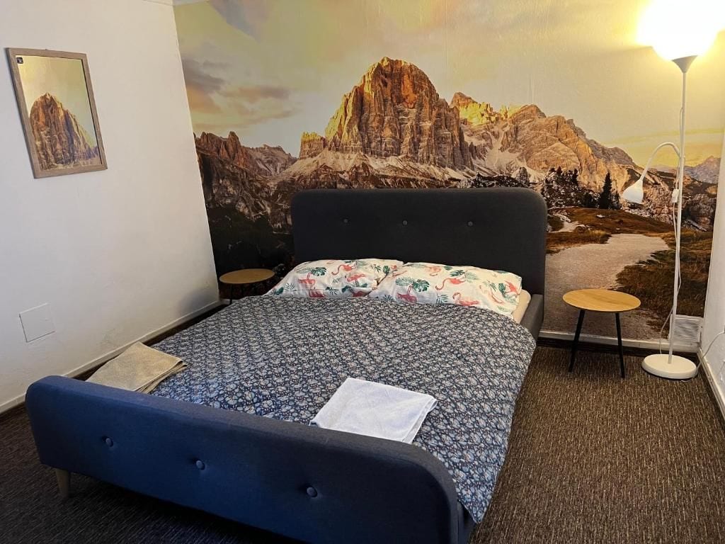 Una habitación de hotel con una cama, una lámpara de pared y un cuadro de un paisaje de montaña.