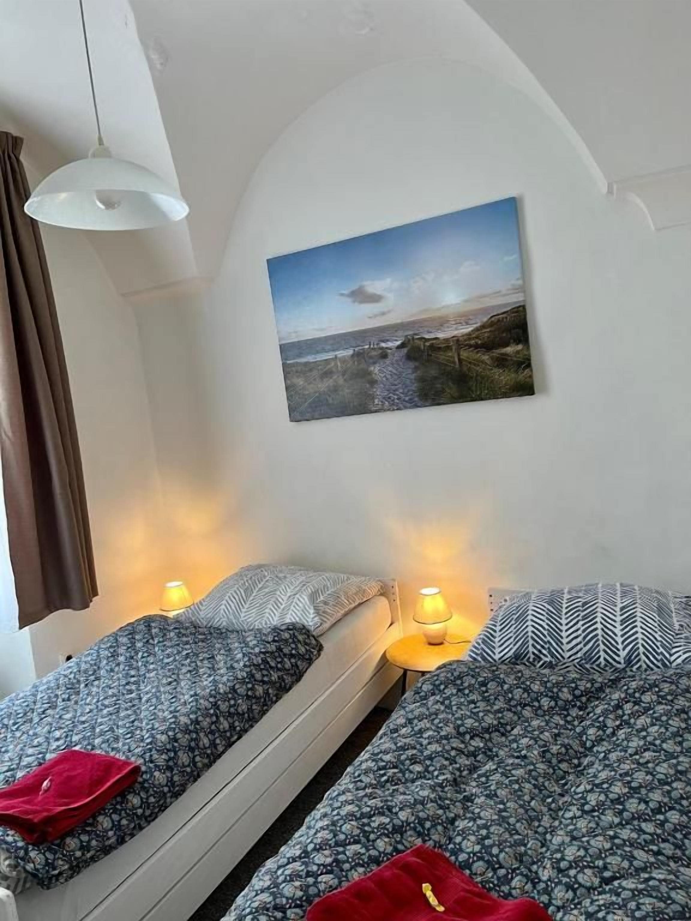 Una habitación con dos camas, un cuadro de un paisaje y una lámpara colgante.