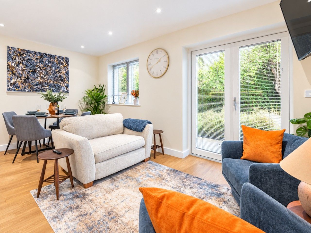 Ein modernes Wohnzimmer mit Sofa, Sesseln, einem Couchtisch und einem Essbereich mit Stühlen und einem Tisch. Ein großes Fenster mit Jalousien, eine Wanduhr und eine Pflanze auf der Fensterbank sind ebenfalls zu sehen.