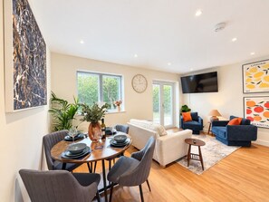 A modern living room with a dining area, a flat-screen TV mounted on the wall, and a large window providing natural light. - Chic 2bed ground floor flat with garden & parking (Cambridgeshire)