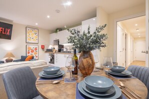 A modern dining area with a wooden table set for four, a vase with greenery, and a kitchen in the background. - Chic 2bed ground floor flat with garden & parking (Cambridgeshire)