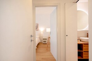A modern hotel room with a wooden floor, a bed with white bedding, a bedside table with a lamp, and a bathroom with a wooden vanity and a round mirror. - Casa Iris – Elegant Modern Retreat in the Heart of Bari (Bari)
