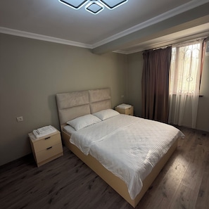 A bedroom with a large bed, bedside tables, a nightstand, a window with curtains, and a ceiling light fixture.