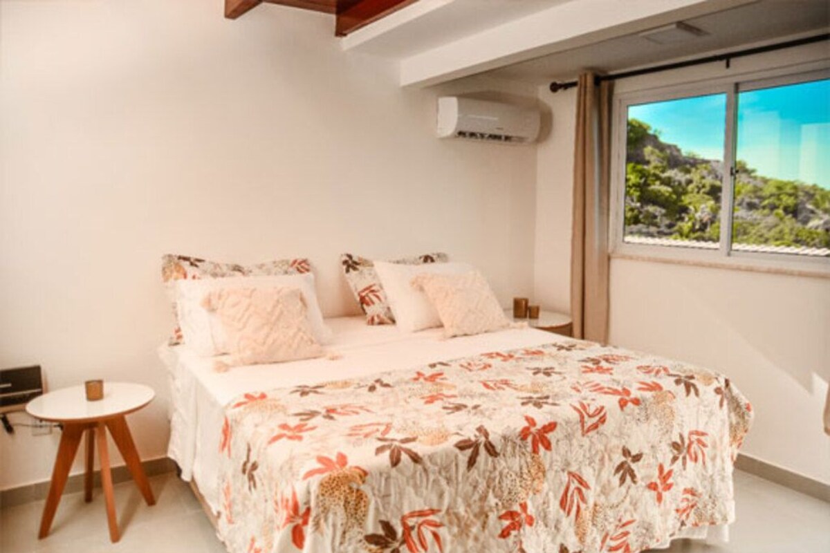 A bedroom with a bed, a small round table, and a window with a view of greenery.
