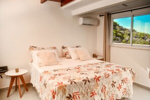 A bedroom with a bed, a small round table, and a window with a view of greenery. - Morro Apartaments (Morro de São Paulo)