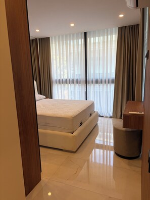 Une chambre à coucher moderne avec un grand lit, une table de chevet et une vue sur l’extérieur à travers des rideaux légers.