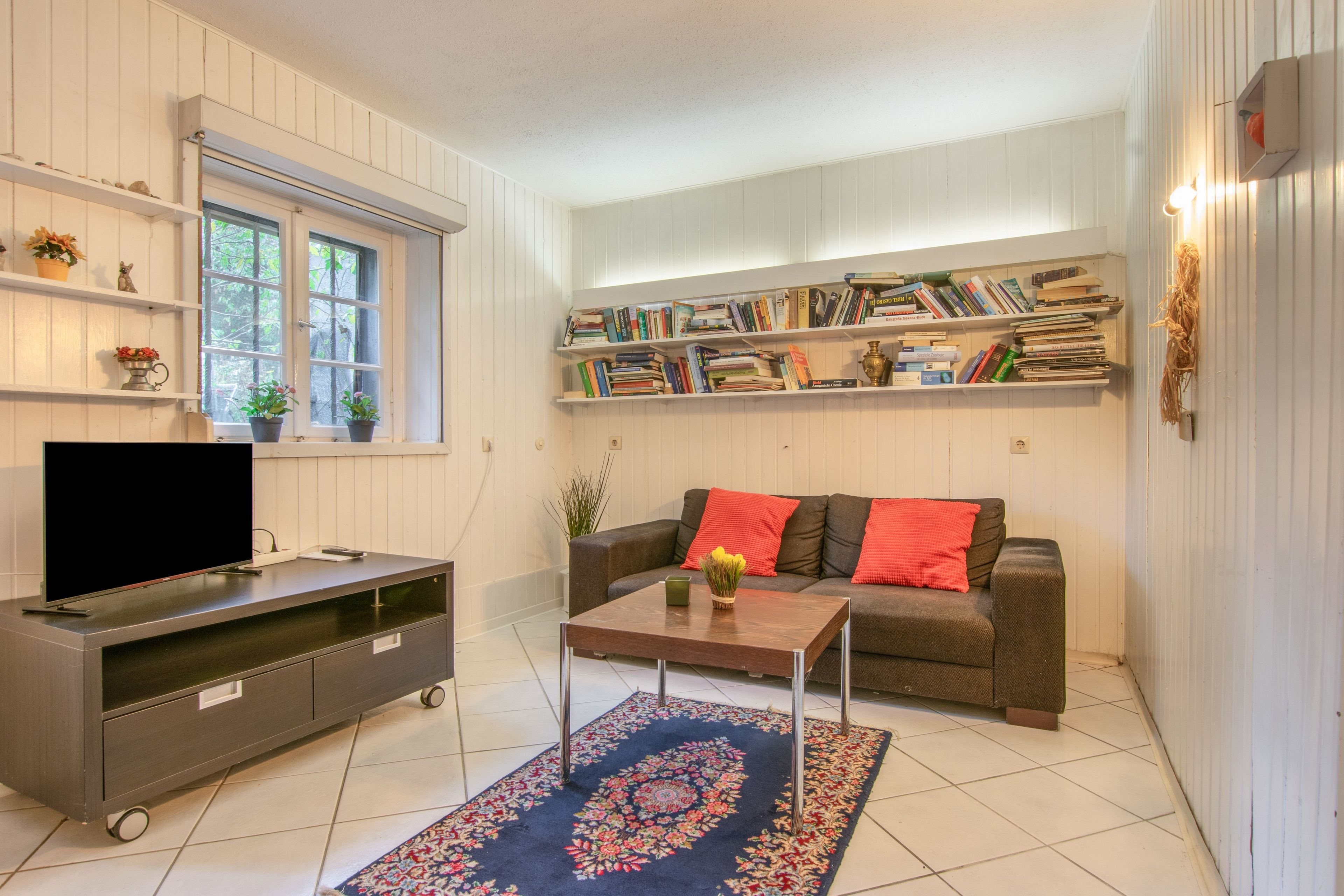 Ein Wohnzimmer mit Flachbildfernseher, einer Couch mit roten Kissen, einem Couchtisch und einer Bücherwand voller Bücher.