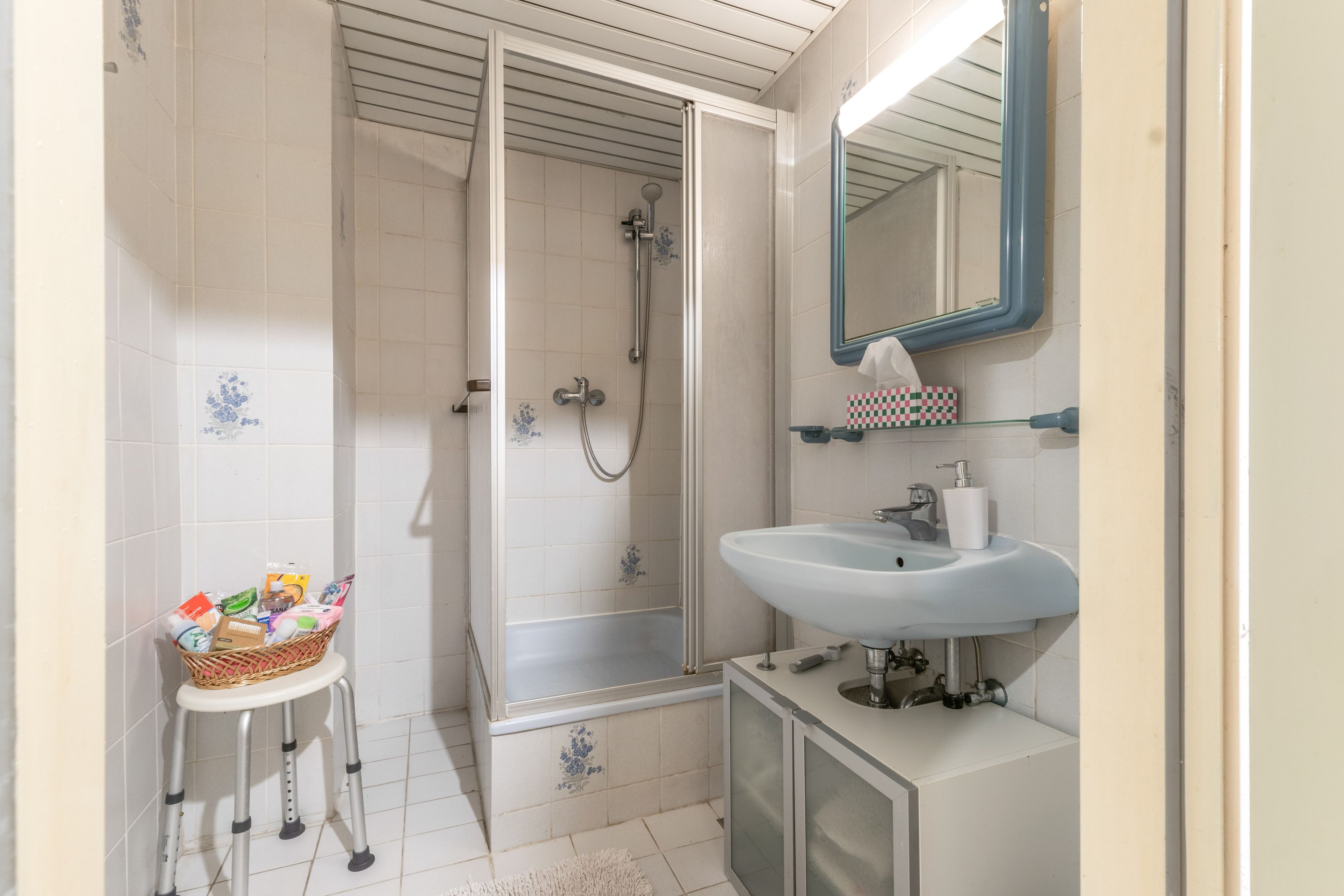 Ein Badezimmer mit Dusche, Waschbecken, Spiegel und einem kleinen Hocker mit Pflegeprodukten.