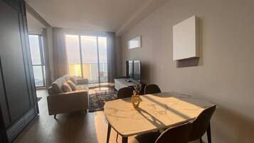 A modern living room with a sofa, a dining table with chairs, a TV, and a balcony with a view.