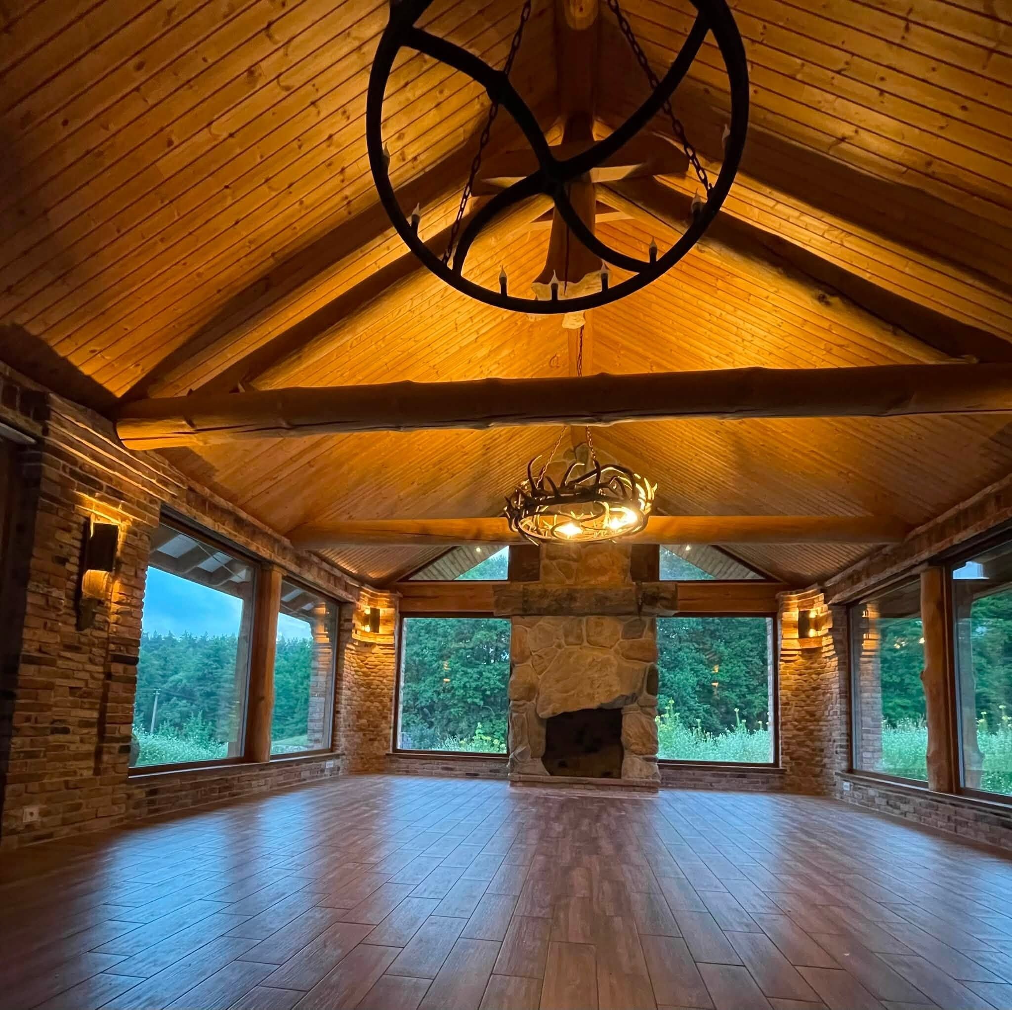 A spacious room with a high ceiling, stone fireplace, and large windows offering a view of the outdoors.