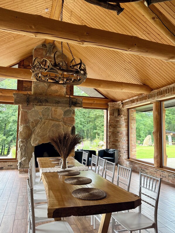 A dining area with a stone fireplace, wooden ceiling, and large windows overlooking a garden.