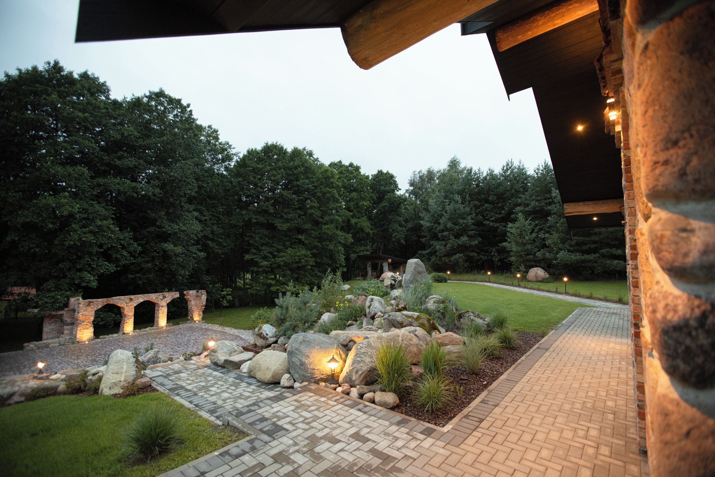 A landscaped garden with a paved pathway, a stone archway, and a well-lit outdoor area.