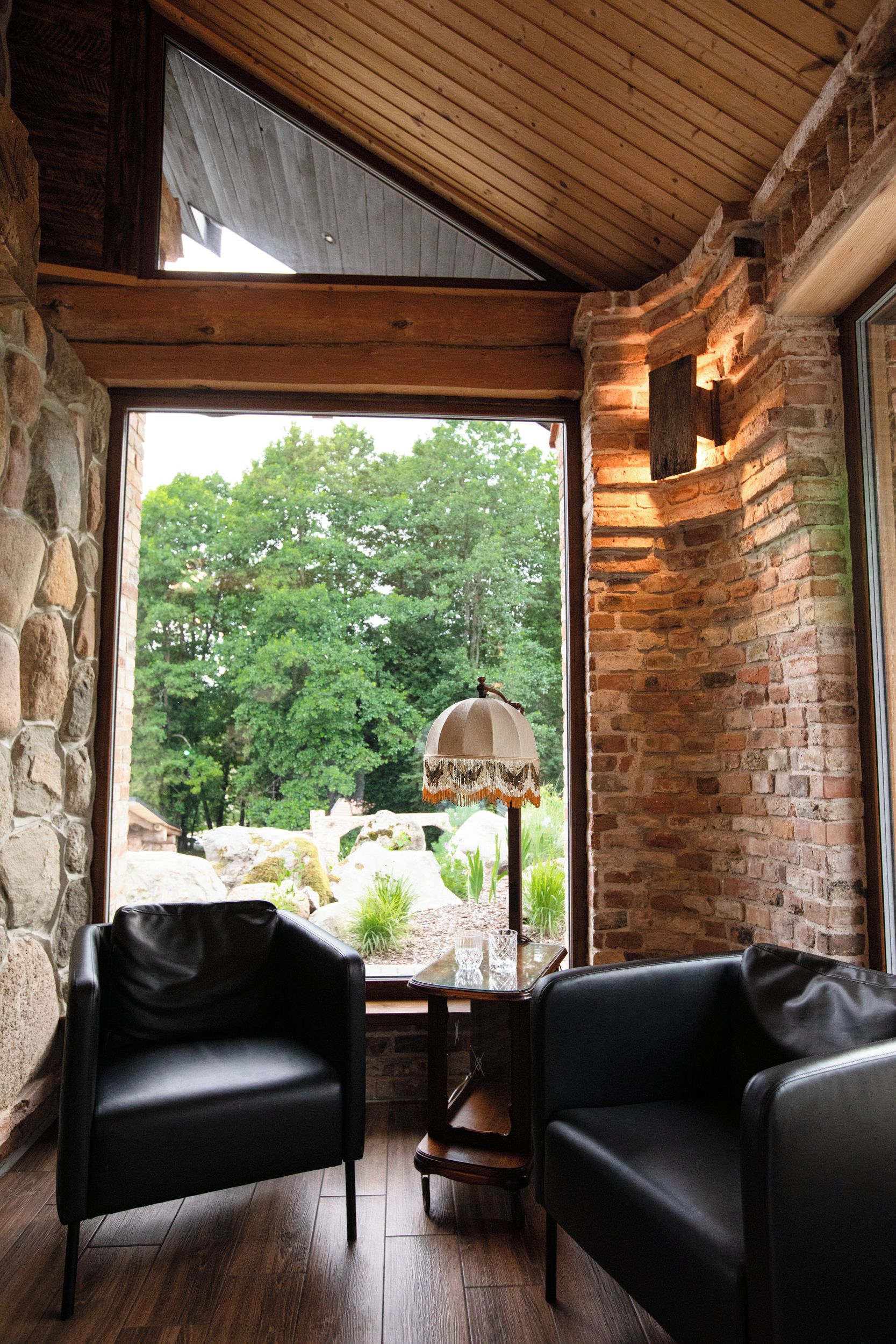 Une pièce avec un mur en pierre, un plafond en bois et une vue sur les arbres par une grande fenêtre. On y trouve deux fauteuils en cuir noir et une petite table avec une lampe.