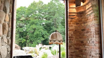 Une pièce avec un mur en pierre, un plafond en bois et une vue sur les arbres par une grande fenêtre. On y trouve deux fauteuils en cuir noir et une petite table avec une lampe.
