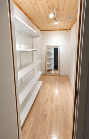A room with wooden flooring, white walls, and a ceiling with exposed beams. There are white shelves on the left wall and a door on the right. - Ovaragøta 60 (Hoyvík)