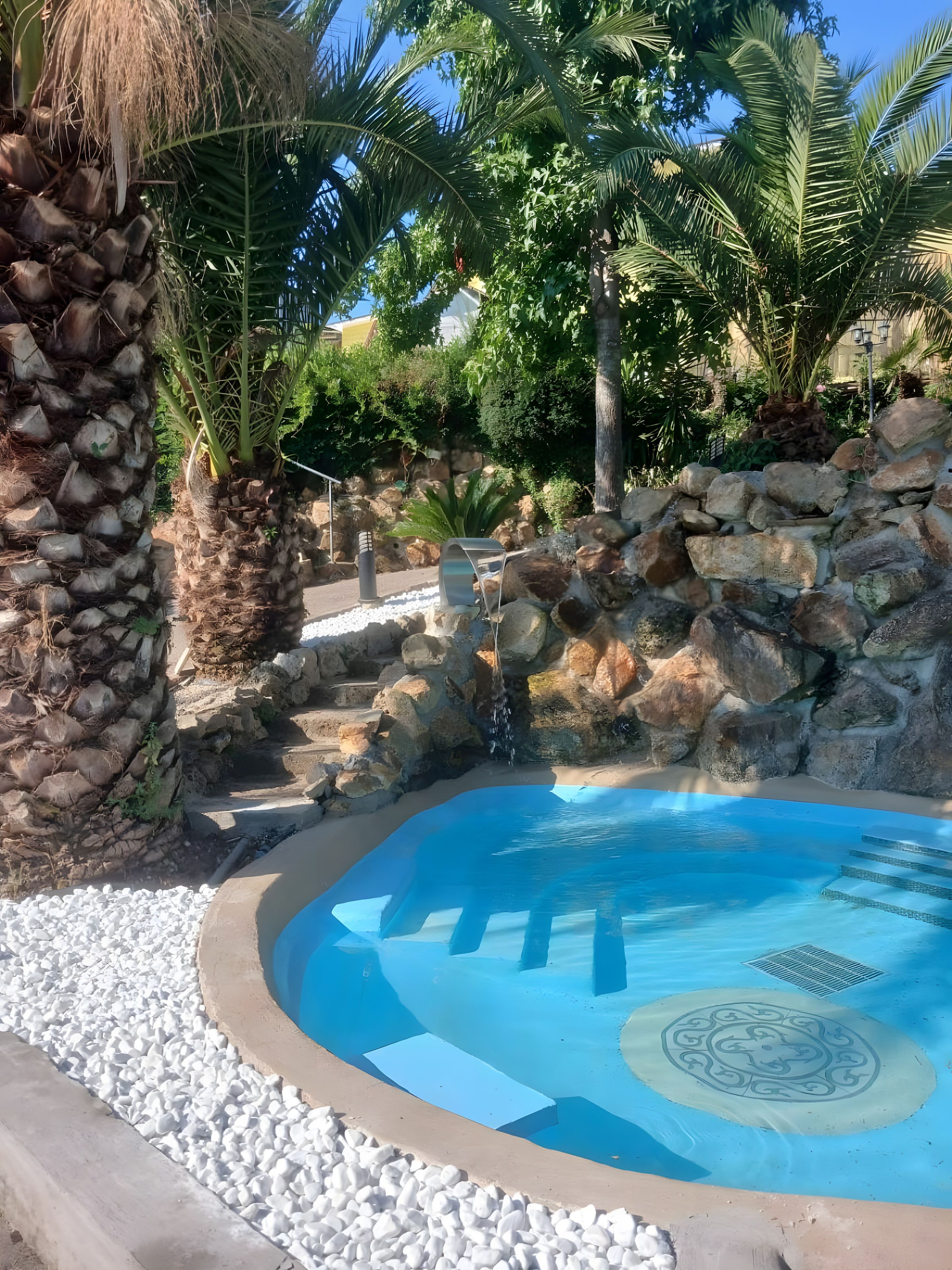 Une piscine à l’eau d’un bleu cristallin, entourée de petits galets blancs, d’un mur de pierre et de palmiers.