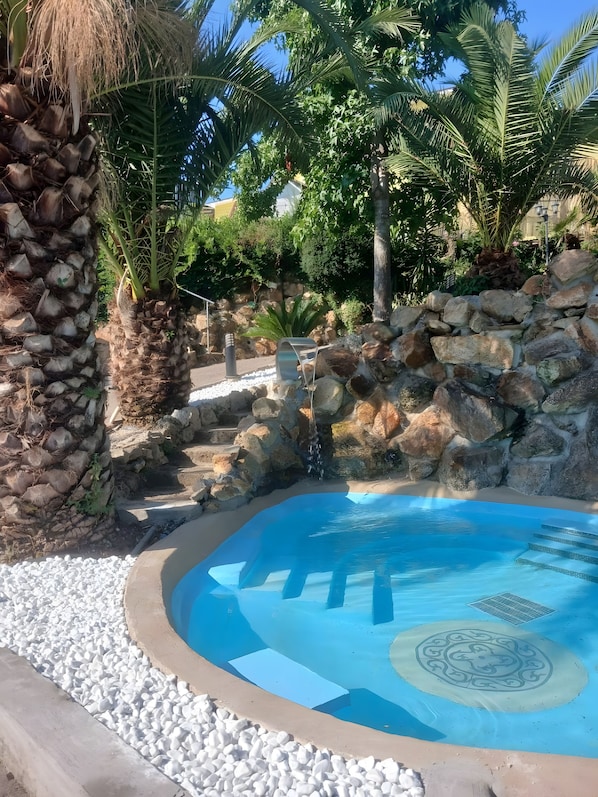 Una piscina con un centro azul cristalino rodeada de piedras blancas, un muro de piedra y palmeras.