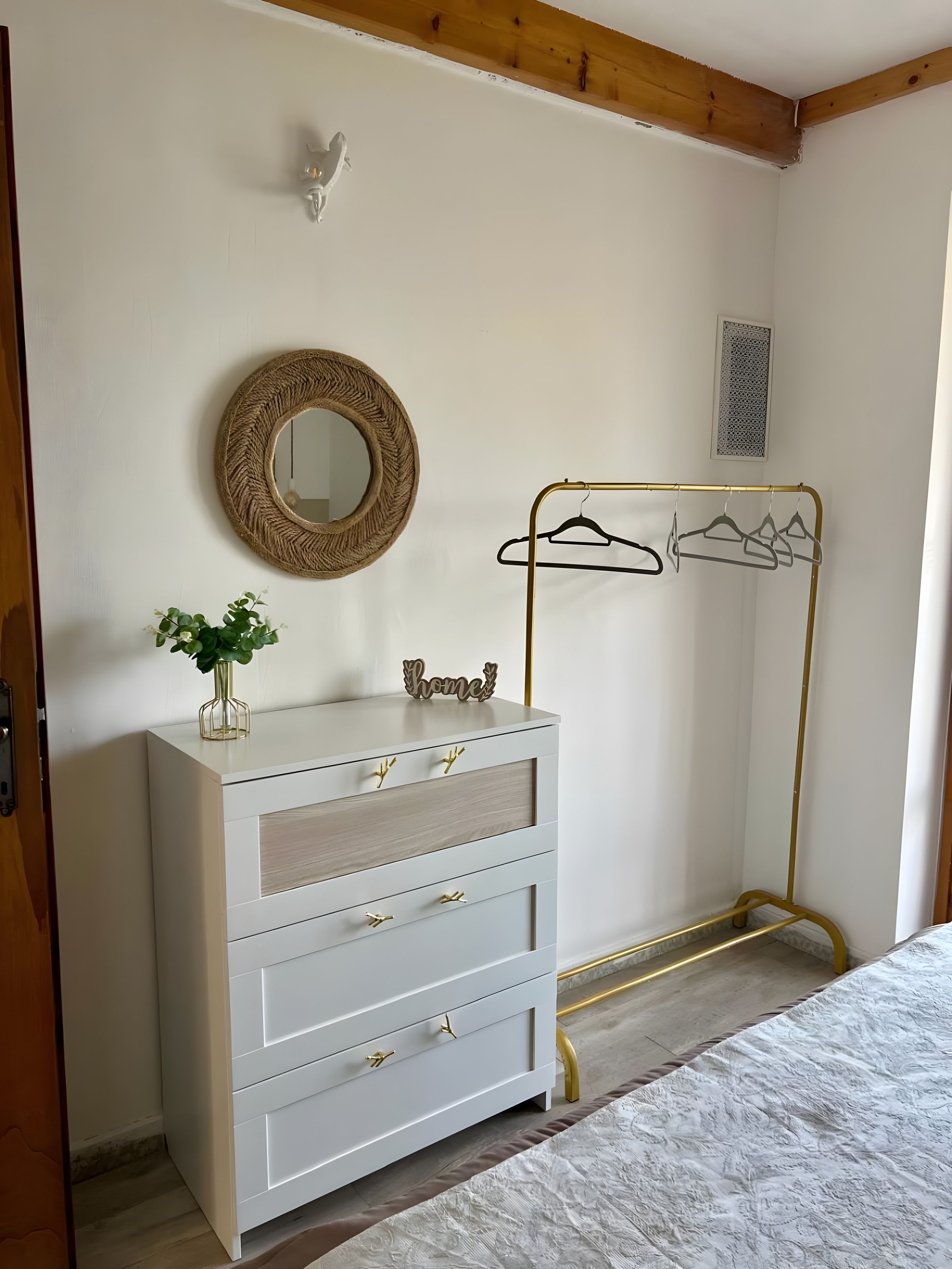 A room with a white dresser, a mirror, a gold hanger rack, and a bed with a textured white bedspread.