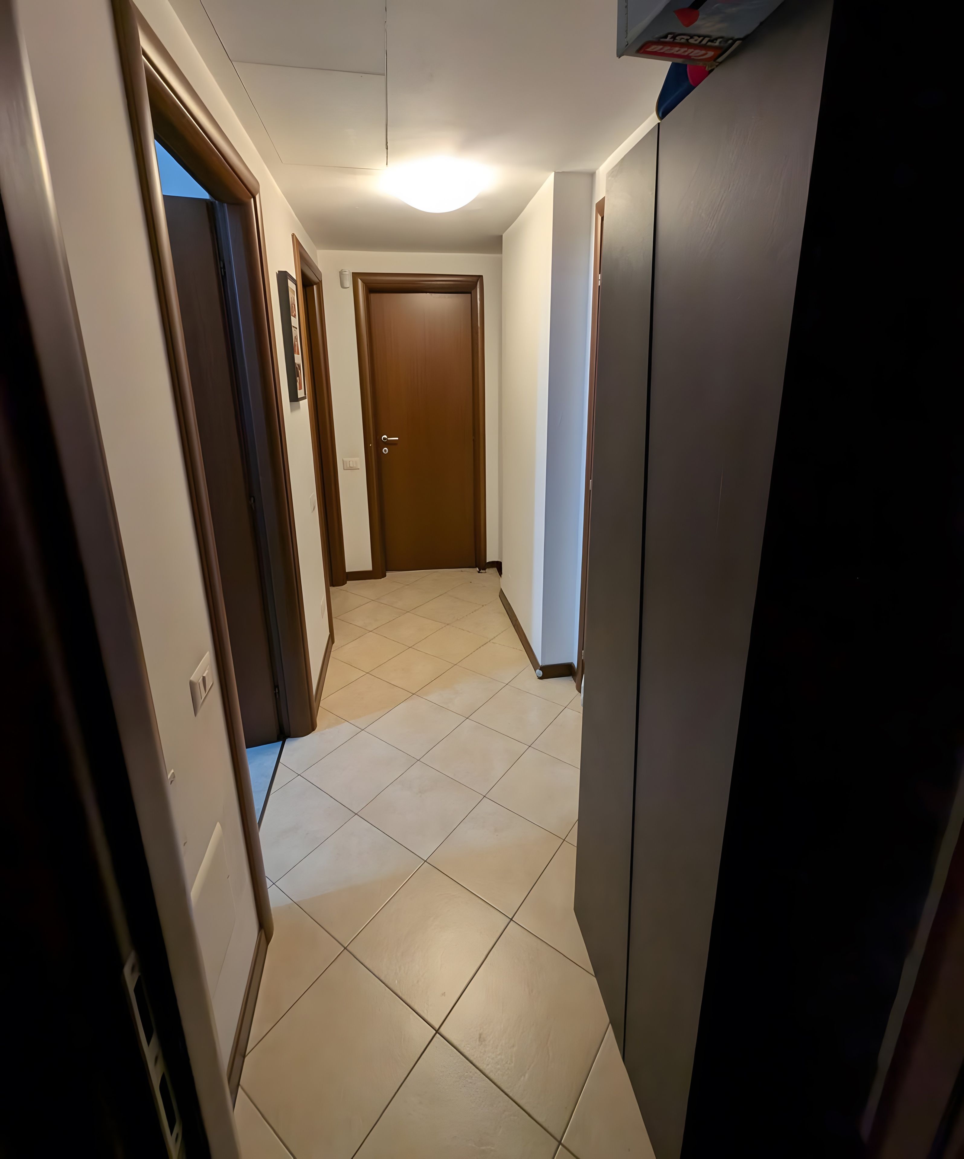 Un couloir avec une porte au fond, un sol carrelé et un luminaire au plafond.