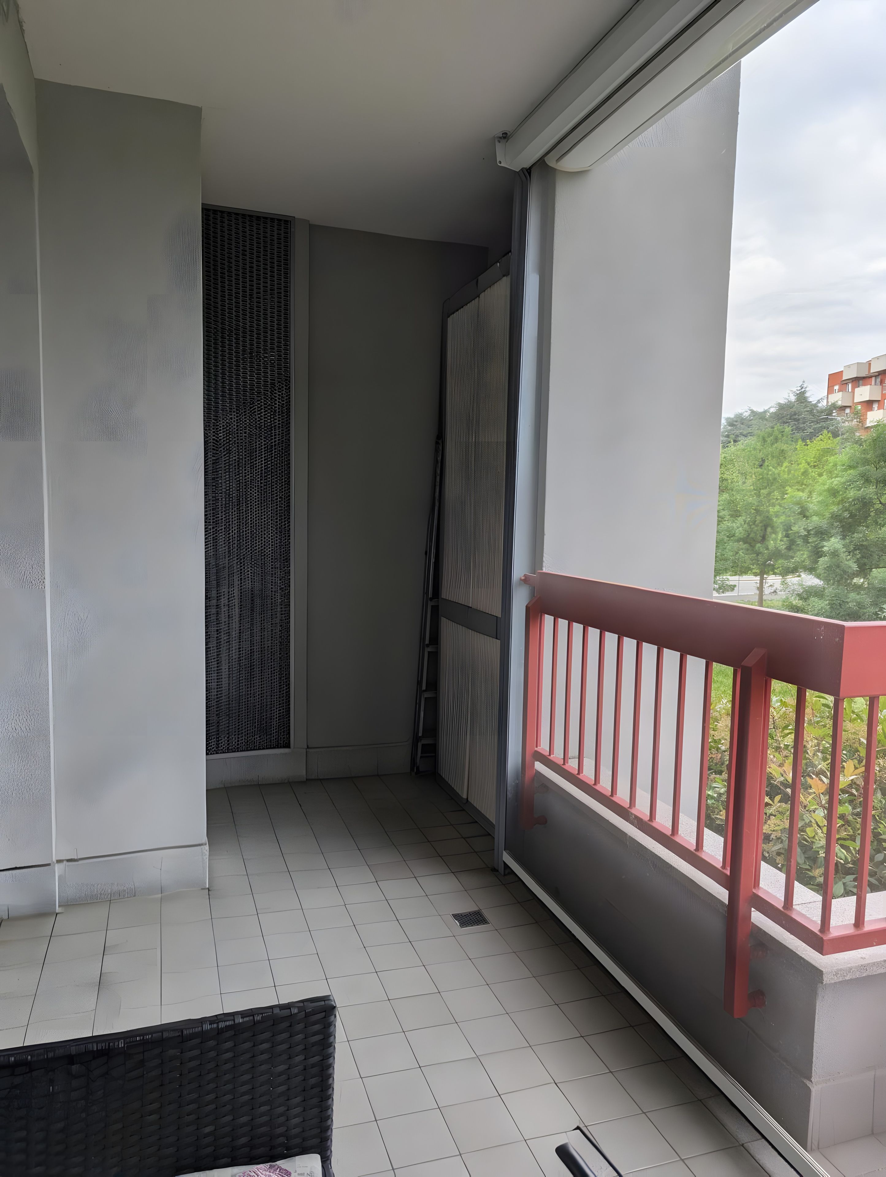 Un balcon avec une balustrade rouge, un sol carrelé et une vue sur la verdure et les bâtiments en arrière-plan.
