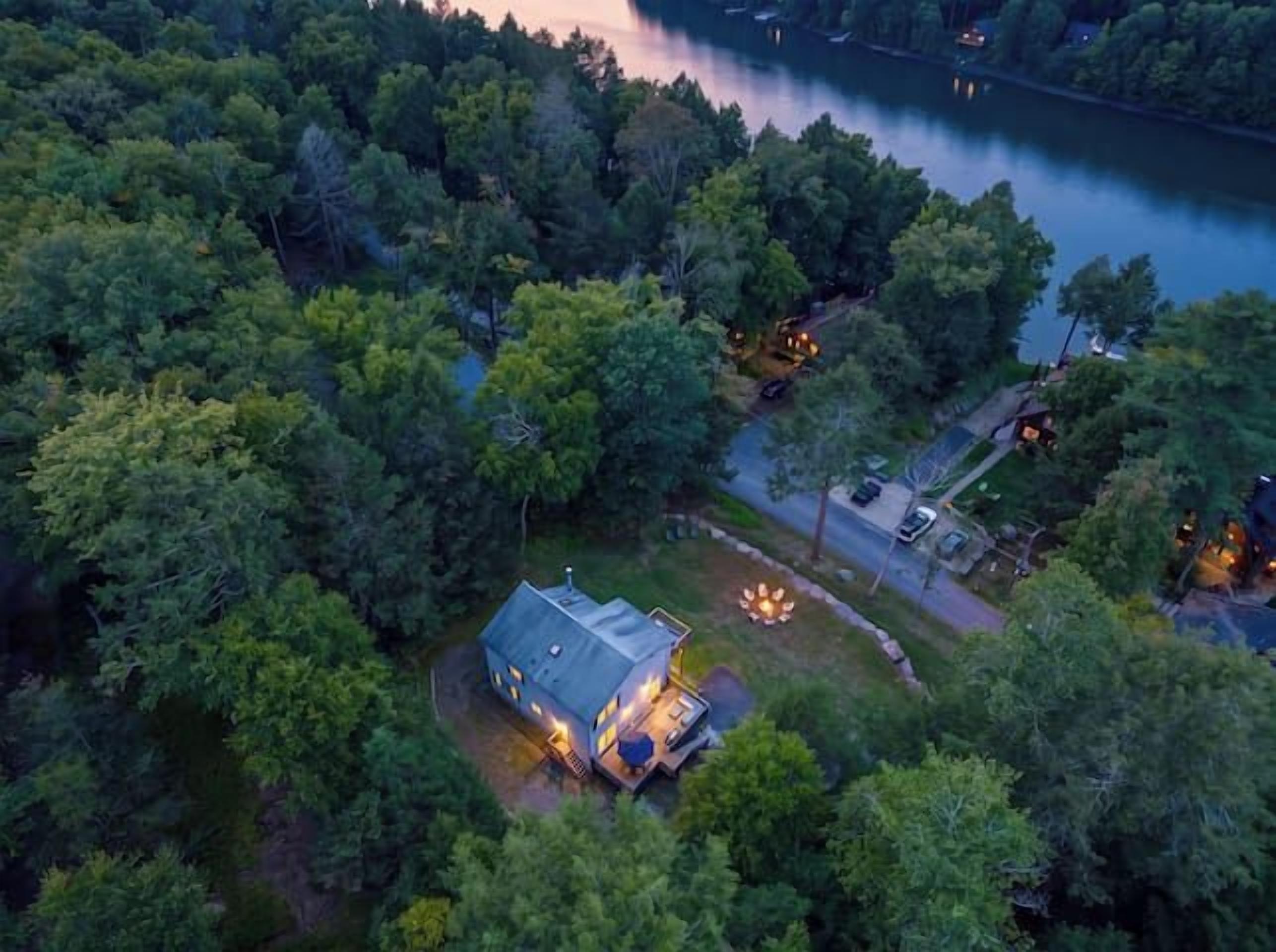 A house surrounded by dense forest with a visible driveway and a lit outdoor area.