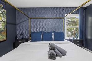 Cama bem arrumada com edredom branco e almofadas azuis, janela com cortinas e um quadro na parede.