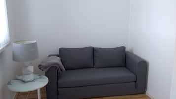A small, neatly arranged room with a grey sofa, a round table with a lamp, and a window.