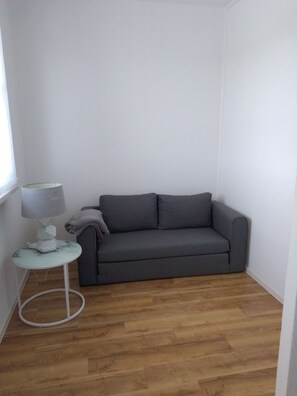 Una habitación pequeña y ordenada con un sofá gris, una mesa redonda con una lámpara y una ventana.