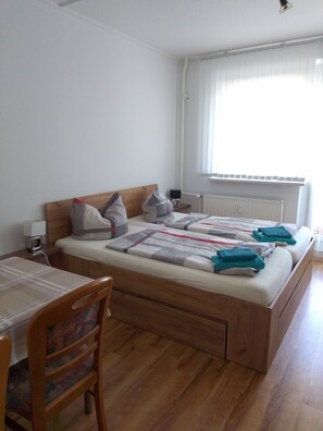 A bedroom with a wooden bed, a bedside table with a lamp, a chair, and a window with blinds.