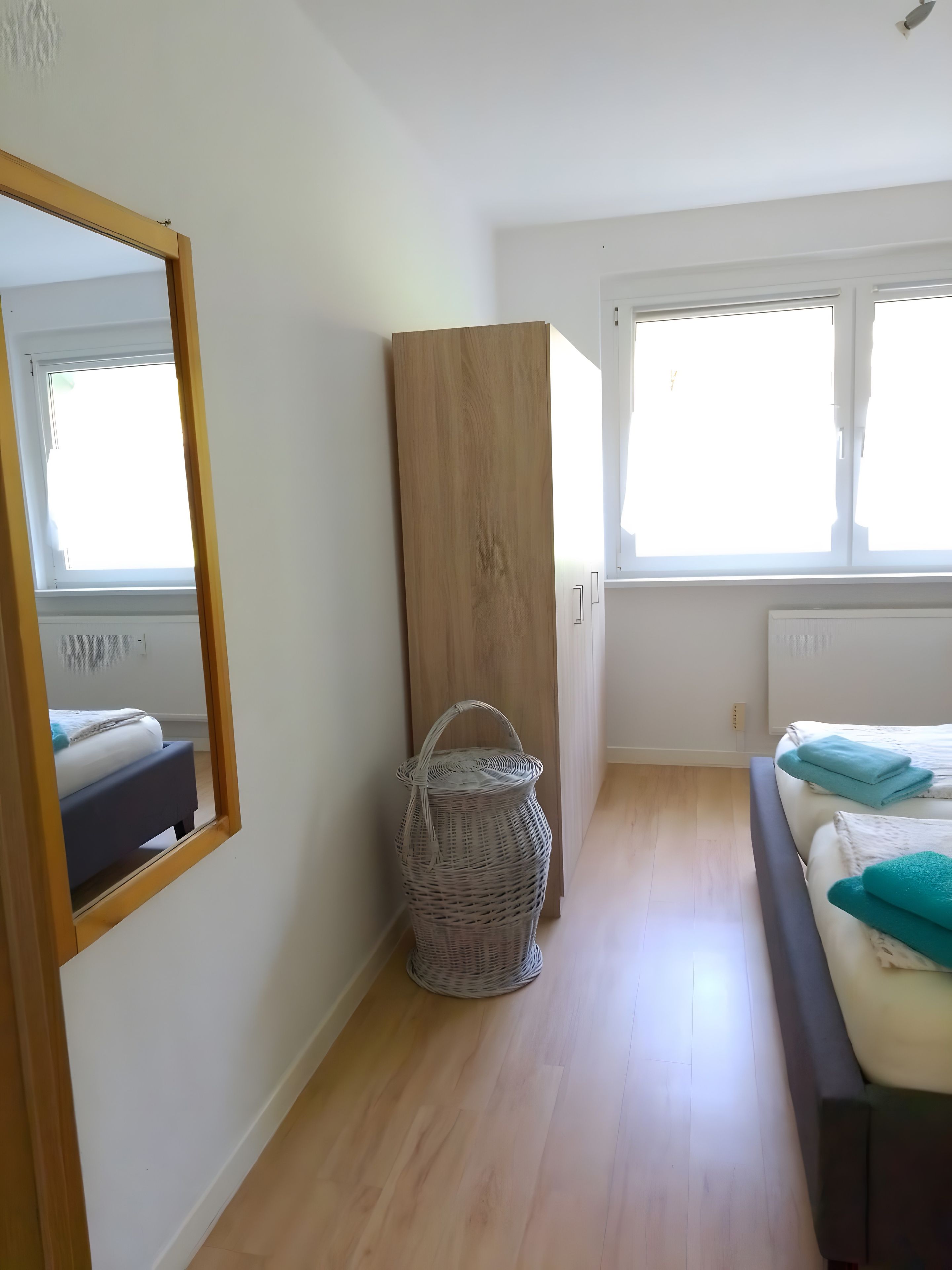 A bedroom with a bed, a wooden wardrobe, a wicker basket, and a large mirror.