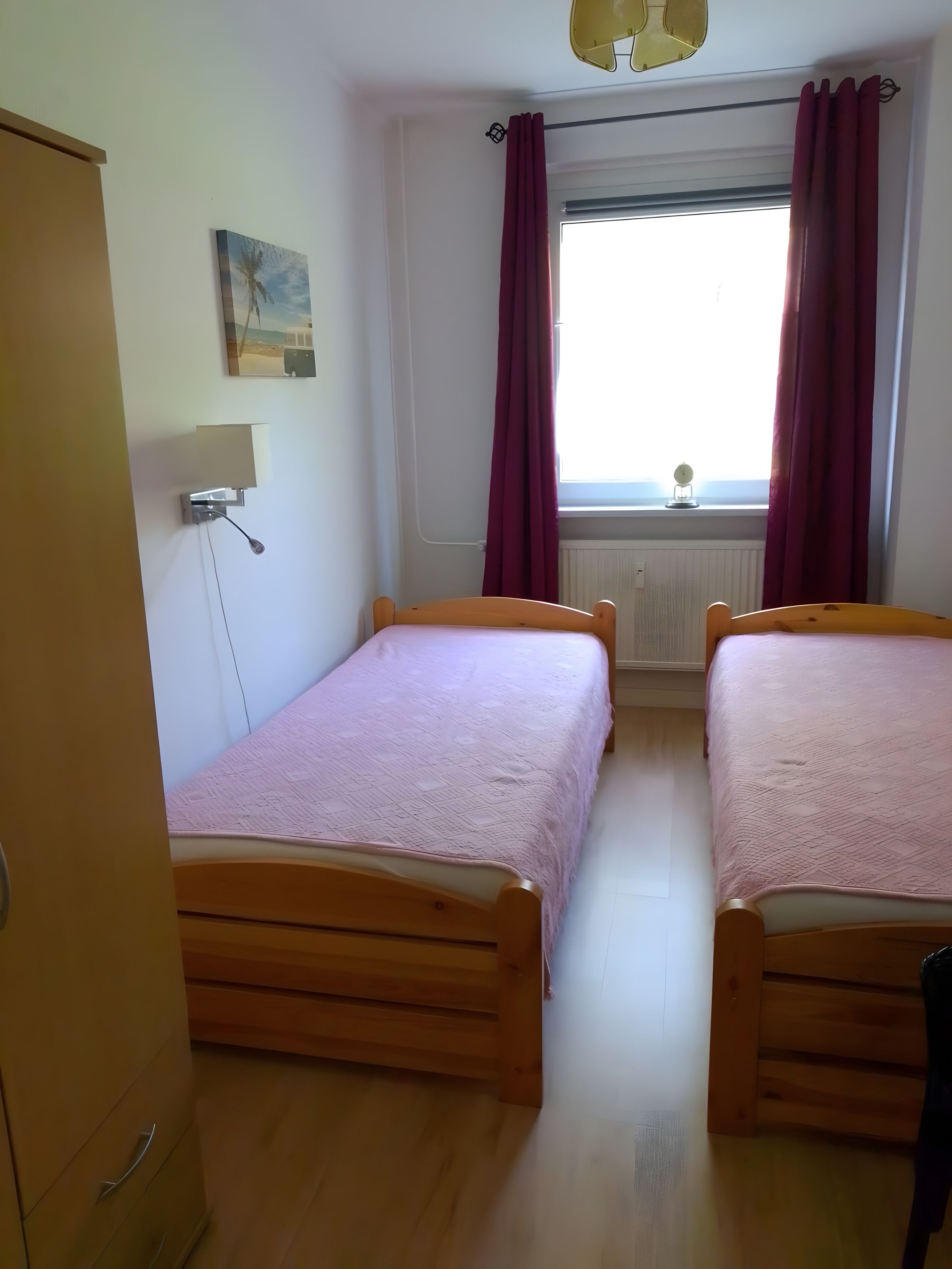 Ein Zimmer mit zwei hölzernen Betten, einer Wandleuchte, einem Fenster mit roten Vorhängen und einer Heizung.
