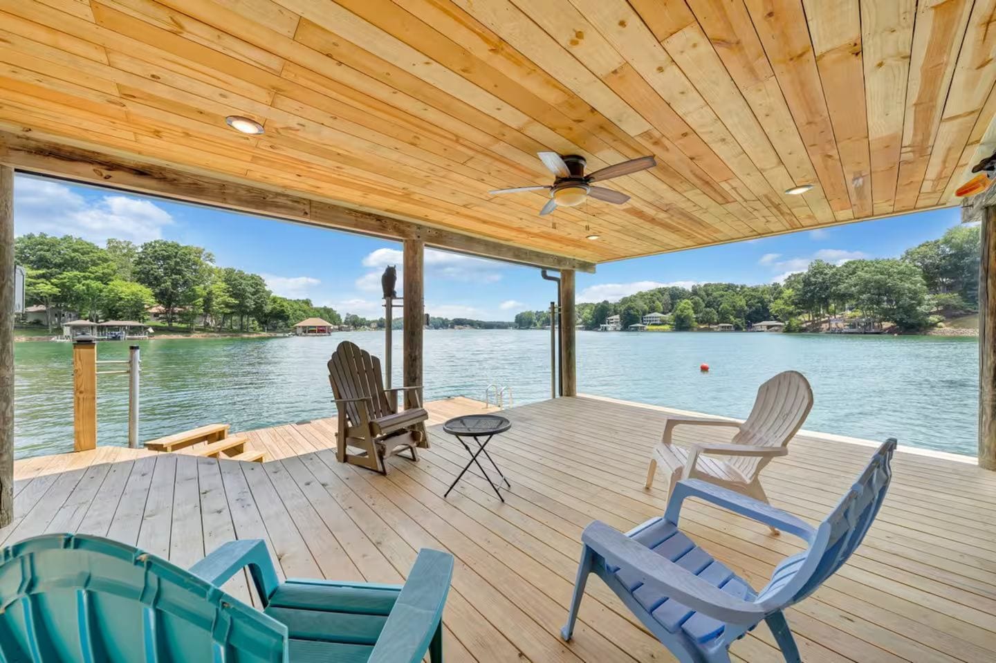 A covered deck with wooden flooring, two Adirondack chairs, a small table, and a ceiling fan.