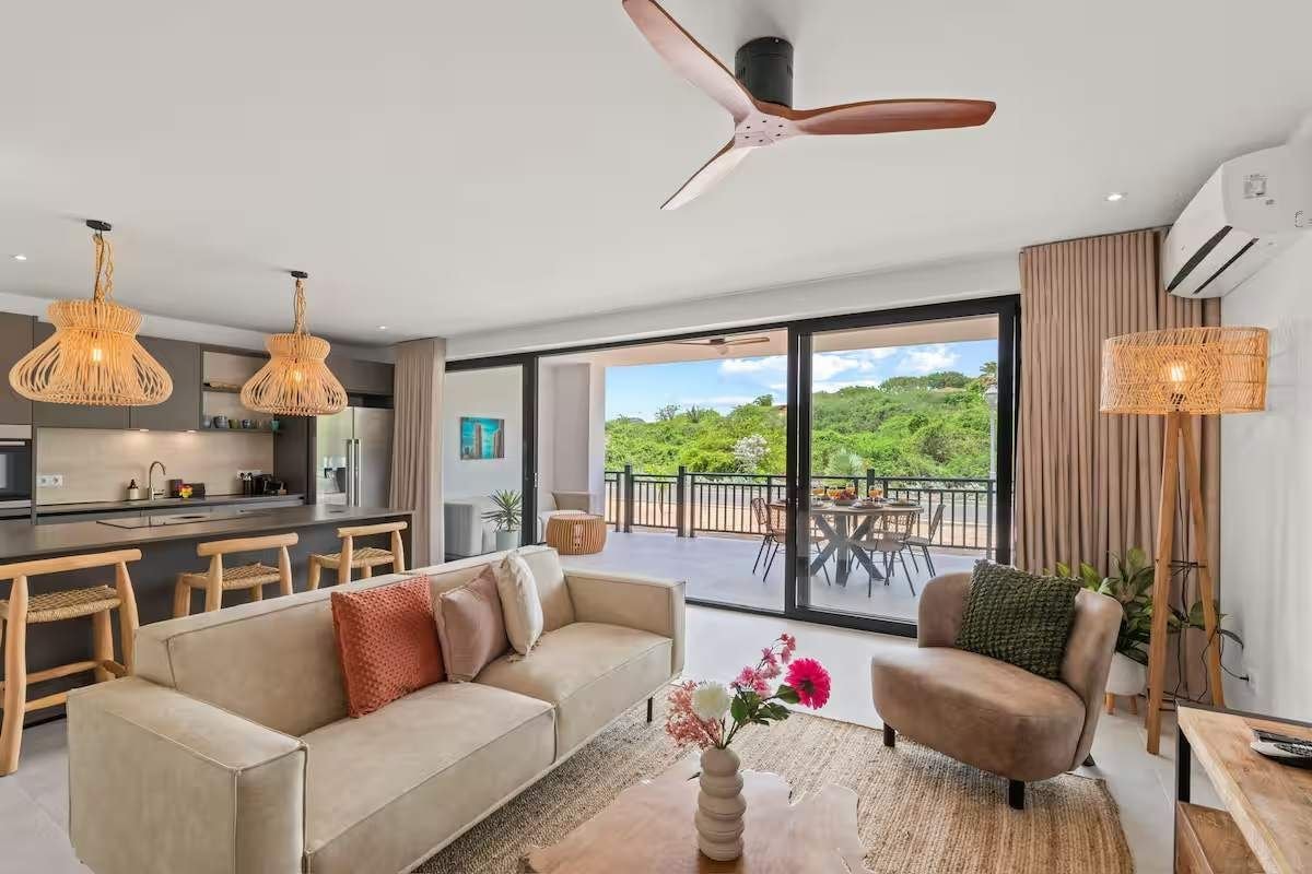 Ein modernes Wohnzimmer mit Sofa, Sessel und Couchtisch, ein Küchenbereich mit Barhocker und ein Balkon mit Gartenmöbeln.