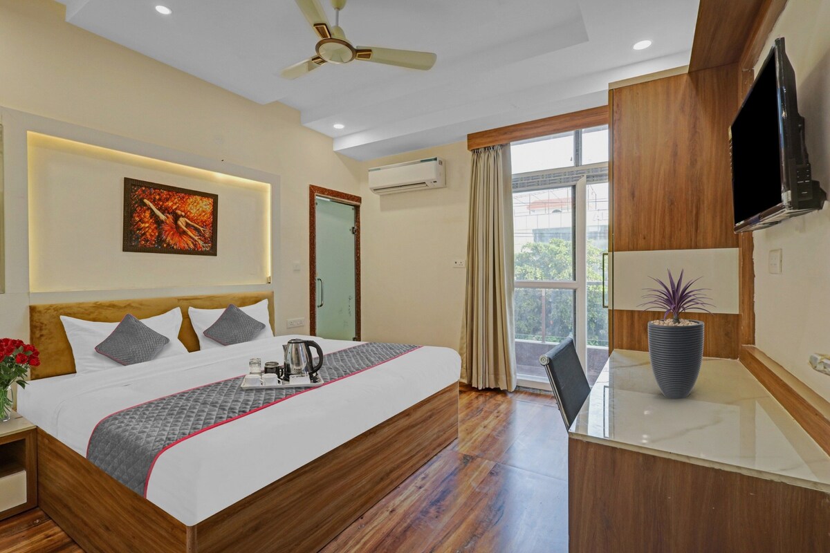 A hotel room with a bed, bedside table, a flat-screen TV, a potted plant, and a window with curtains.