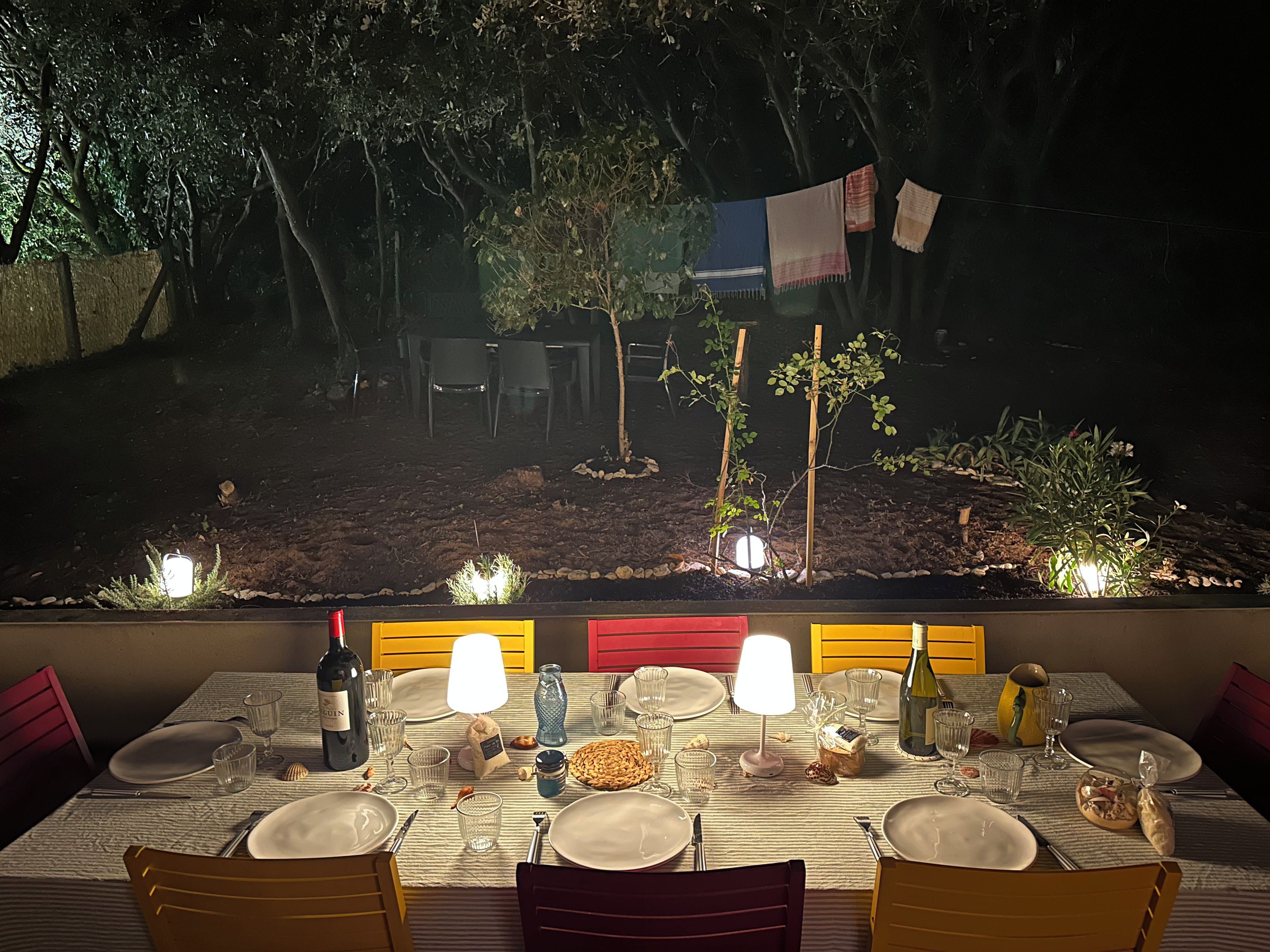Un área de comedor al aire libre con una mesa preparada para dos personas, rodeada de sillas y vista a un jardín con árboles y un tendedero.