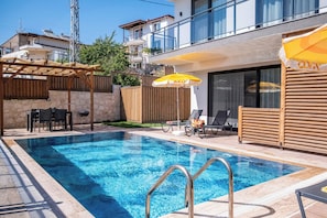 A swimming pool with a wooden deck, lounge chairs, and a covered seating area. - Villa Savaya 1 Antalya (Kaş)