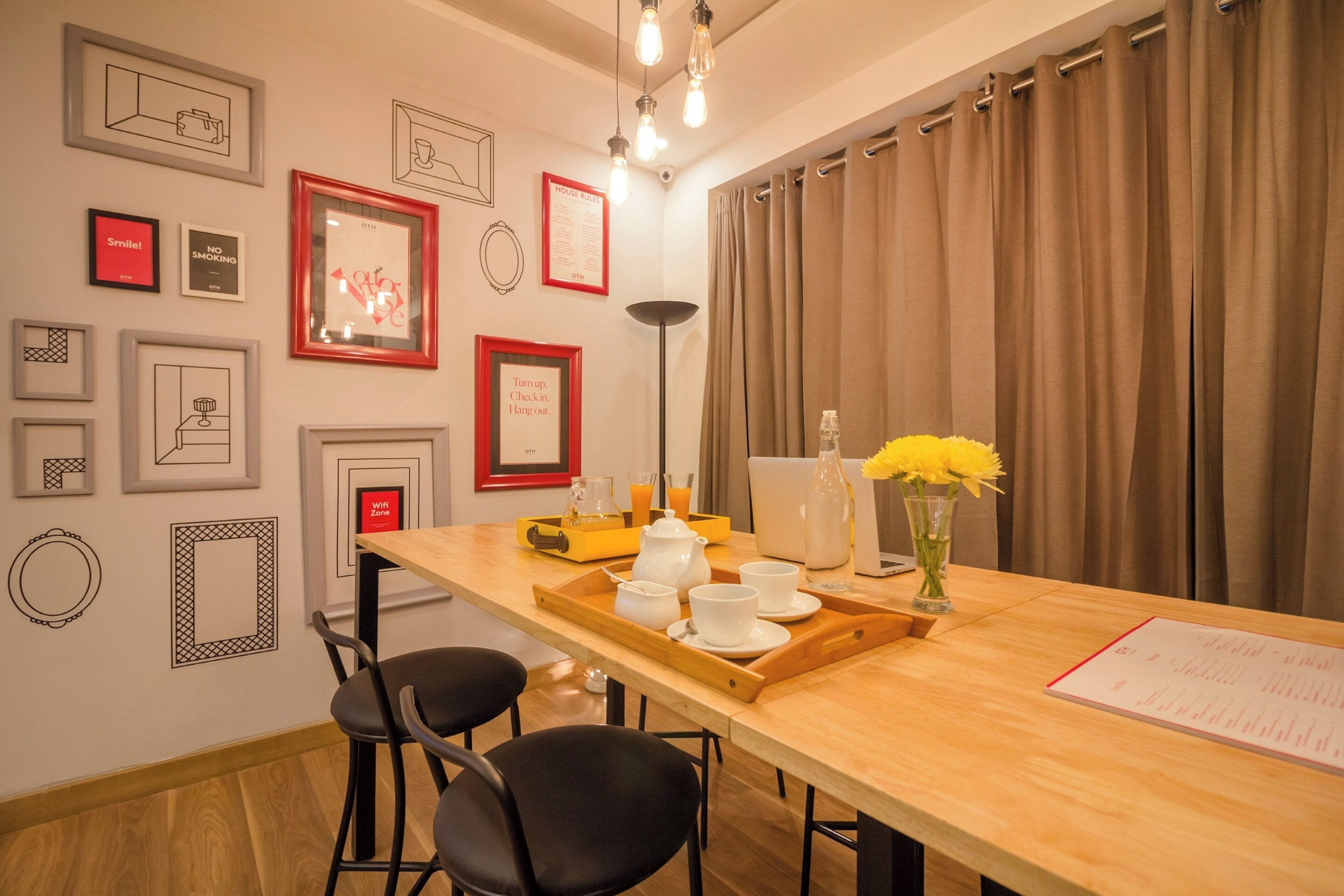 Habitación de hotel con una mesa de comedor de madera, dos sillas negras, una computadora portátil, un jarrón con flores amarillas y varias fotografías enmarcadas en la pared.