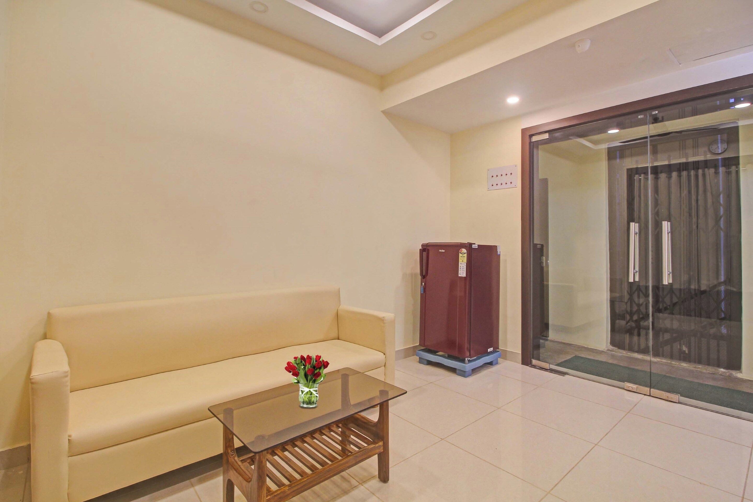 Habitación de hotel con sofá color beige, una mesita de vidrio con un jarrón de flores, un mueble rojo y un vidrio que da a un balcón.