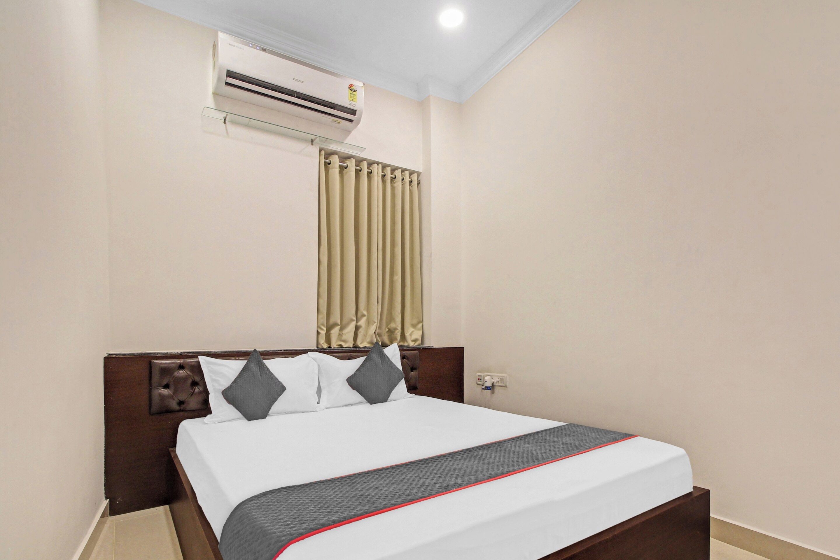 Habitación de hotel con cama, cabecera, ventana con cortinas, unidad de aire acondicionado y un enchufe de pared.