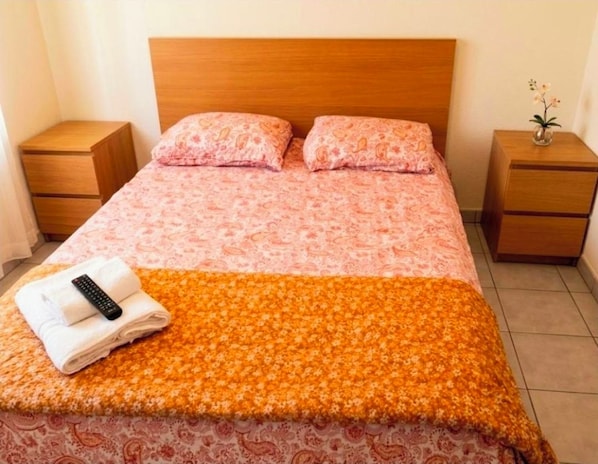 Een net opgemaakt bed met een bedsprei met patroon, twee kussens en een gevouwen handdoek met een afstandsbediening erop. Er staan twee houten nachtkastjes, waarvan er één een vaas met bloemen bevat.