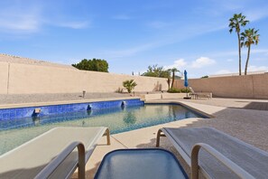 A swimming pool with lounge chairs and a blue-tiled edge, surrounded by a sandy wall and palm trees. - Amazing Desert Retreat w/Saltwater Pool! (Phoenix)