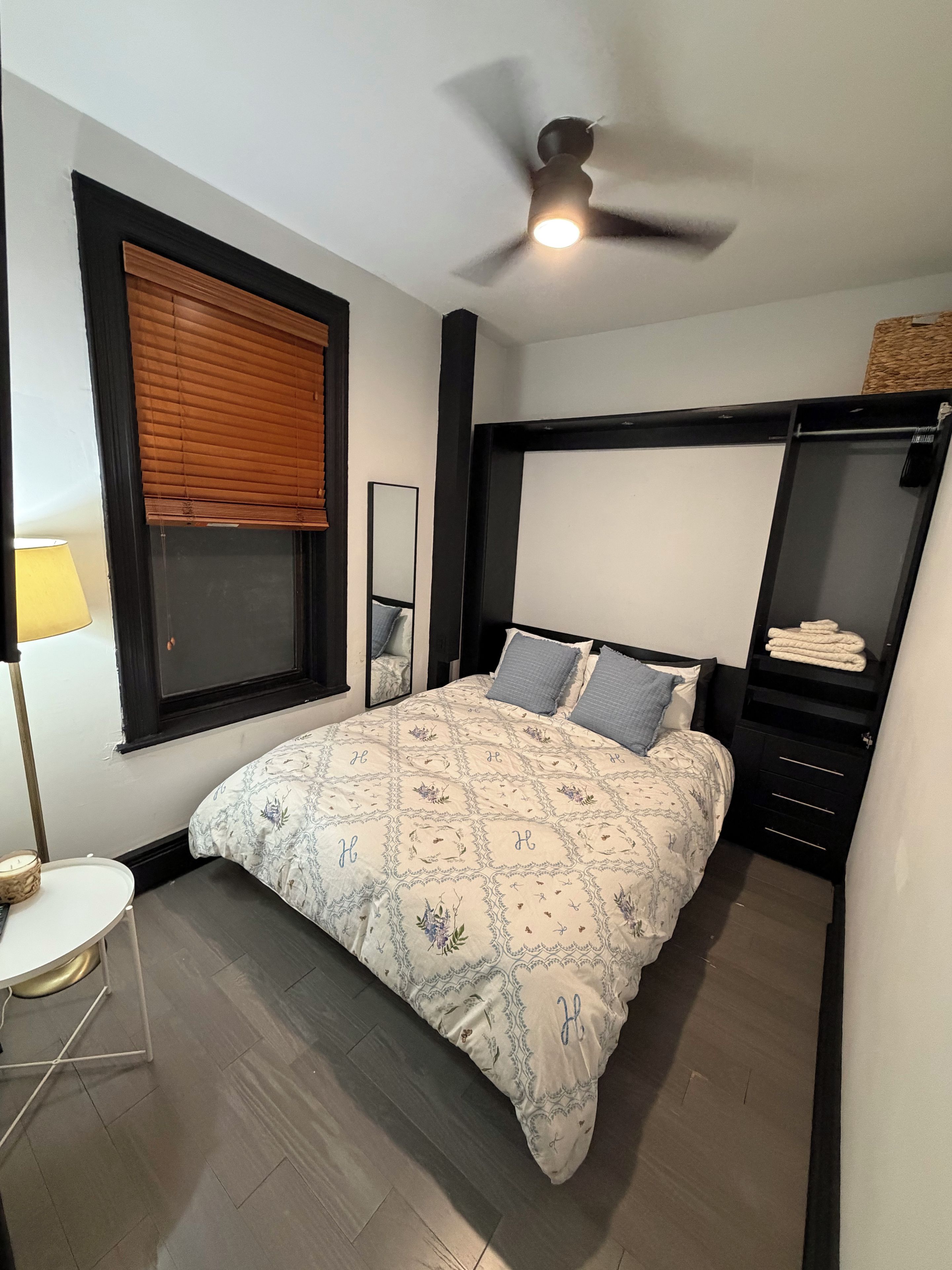 Ein Schlafzimmer mit einem Bett, einem Deckenventilator, einem Fenster mit Jalousien, einem Nachttisch und einem Spiegel.