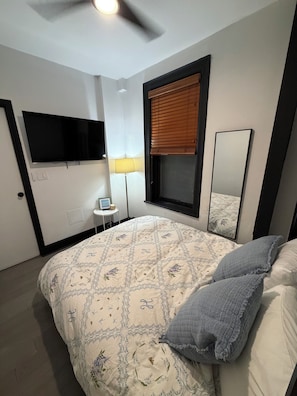 A bedroom with a bed, a flat-screen TV, a bedside lamp, a window with blinds, and a small table with a lamp. - Perfect 1-bedroom apartment in peaceful Hoboken (Hoboken)