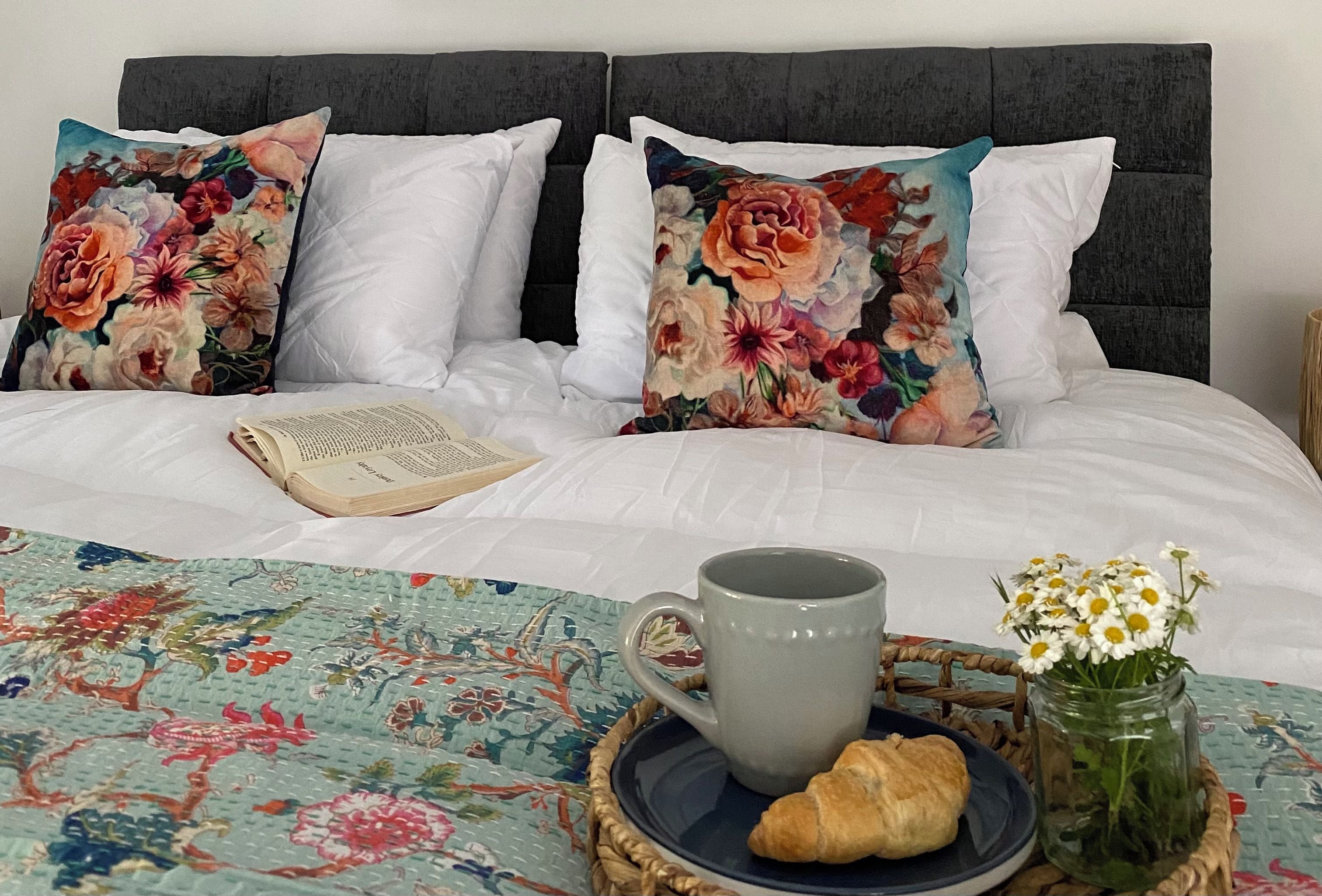 Una cama con almohadones florales, un libro, una taza, un croissant y un florero sobre una mesa.