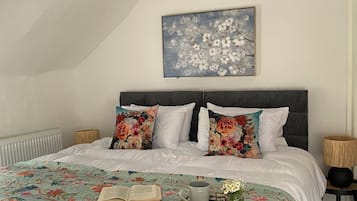 Quarto com uma cama grande, roupa de cama floral, um livro, uma caneca e um cesto com flores.